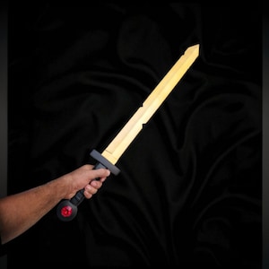 May include: A wooden toy sword with a light yellow blade and a black handle. The handle has a red button. The sword is held in a person's hand against a black background.