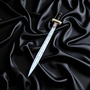 May include: A decorative sword with a silver blade and a brown and gold hilt. The blade has a decorative design along the edge. The sword is set against a black satin background.