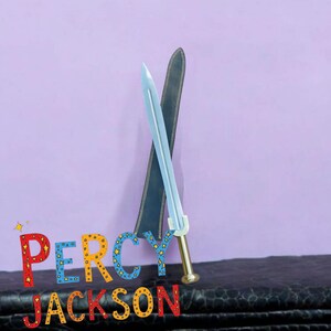 May include: A light blue toy sword with a gold handle and a black sheath, set against a purple background. The text "PERCY JACKSON" is displayed in colorful letters below the sword.