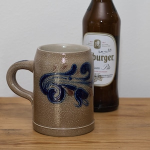 Op de afbeelding: Een bruin keramische bierpul met een handvat, met een blauw en bruin bloemmotief. Op de achtergrond een bruine bierfles met een wit etiket. De pul staat op een houten oppervlak.