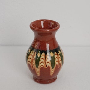 Puede incluir: Un pequeño jarrón de cerámica de color terracota, con cuerpo redondeado y cuello estrecho. El jarrón está adornado con una banda decorativa con un patrón ondulado en verde, amarillo y blanco sobre fondo negro. El jarrón mide unos 10 cm de alto.