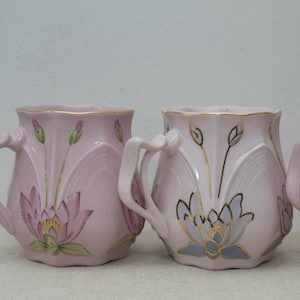 May include: Three pink ceramic mugs with floral designs. The mugs have gold accents and are decorated with pink and blue flowers.