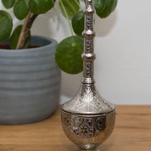 May include: An intricately designed silver container with a tall, decorative stopper. The container features a bulbous base and detailed floral patterns. A potted plant with green leaves is in the background, set on a wooden surface.