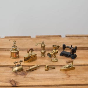 Può includere: Una collezione di miniature in ottone di oggetti per la casa, tra cui un ferro da stiro, una macchina da cucire e un mattarello, disposti su una superficie di legno. Gli oggetti sono dettagliati e sembrano essere vintage o antichi. La tavolozza dei colori è principalmente oro e marrone.