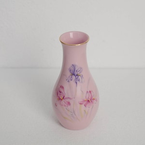 May include: A pink ceramic vase with a gold rim. The vase is decorated with painted iris flowers in shades of purple and pink. The vase is a decorative item.