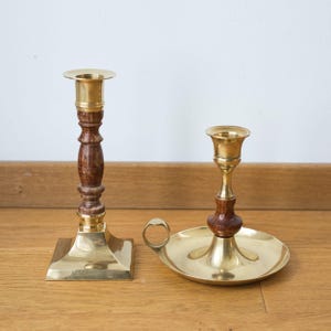 Può includere: Due candelabri vintage in ottone. Uno ha una base quadrata e un centro in legno. L'altro ha una base rotonda con un manico e un centro in legno. Entrambi hanno una finitura dorata e sono progettati per contenere candele.