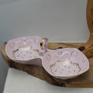 May include: A light pink, two-sectioned ceramic serving dish with a gold handle. The dish has gold trim and a floral design in the centre of each section. The dish is placed on a wooden surface.