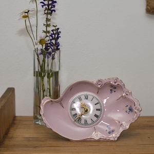 May include: A pink ceramic clock in a shell shape with a white clock face and black Roman numerals. The clock has gold trim and blue floral accents. A clear glass vase with flowers sits behind the clock.