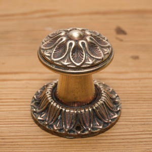 Puede incluir: Un pomo de puerta de latón antiguo con un diseño floral ornamentado. El pomo presenta una parte superior redondeada con un detalle central y una base decorativa. El latón tiene una pátina cálida y envejecida, lo que sugiere su antigüedad. El pomo está diseñado para la instalación en una puerta.