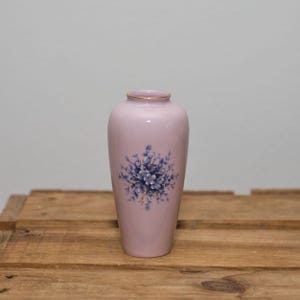 May include: A small, pale purple vase with a gold rim and a floral design. The vase features a cluster of blue flowers with green leaves. The vase is sitting on a wooden surface.