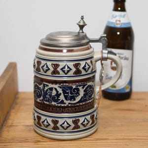 Peut inclure: Une chope à bière en céramique avec un couvercle en étain à charnière. La chope présente une base crème avec des bandes décoratives marron et bleu. Une bouteille de bière est en arrière-plan.