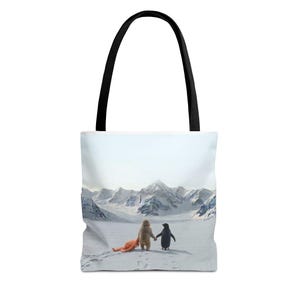 Può includere: Borsa tote bianca con manici neri. Raffigura una scena di montagna innevata con un orso e un pinguino che si tengono per mano. Una figura arancione giace nella neve.
