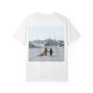 Può includere: T-shirt bianca con una grafica di un orso e un pinguino che si tengono per mano in un paesaggio innevato con montagne sullo sfondo. Una terza figura è sdraiata a terra. L'immagine ha uno stile stravagante e artistico.
