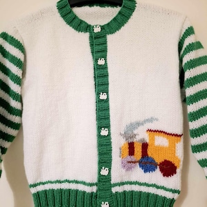Pode incluir: Cardigã branco de malha com mangas e acabamentos listrados verdes. O cardigã apresenta gola e carcela de botões verdes, com botões brancos. Um design de trem colorido está na frente, com detalhes amarelos, azuis, vermelhos e roxos.