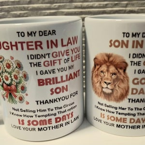 May include: Two white ceramic mugs with black and red text. The left mug features a bouquet of daisies and text addressed to a daughter-in-law. The right mug displays a lion portrait and text addressed to a son-in-law, both signed by a mother-in-law.