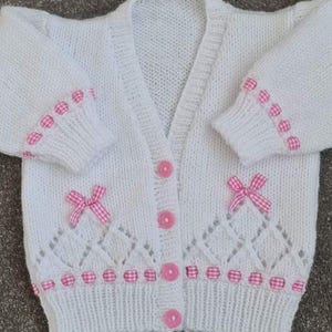 May include: A white knitted baby cardigan with pink detailing. The cardigan has pink bows, buttons, and trim. The sleeves and hem are decorated with pink gingham ribbon. The front features a diamond-shaped openwork pattern.