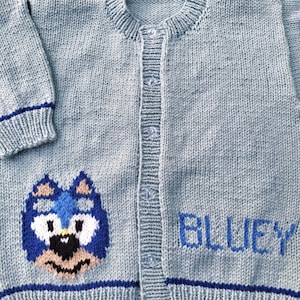 May include: A light blue knitted cardigan with a round collar and button closure. The cardigan features a cartoon dog face and the name "BLUEY" embroidered in blue. The cuffs and hem are trimmed with a dark blue stripe.
