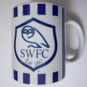 May include: White ceramic mug with a blue and white design. The mug features a blue shield with an owl graphic and the letters "SWFC" with "Est. 1867" below. The mug has a white handle and blue vertical stripes.