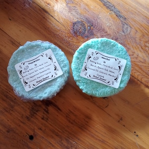 Puede incluir: Dos pastillas de ducha redondas de color azul claro con etiquetas blancas sobre una superficie de madera. Las etiquetas enumeran ingredientes como bicarbonato de sodio y aceites esenciales. Una está etiquetada como "Peppermint" y la otra como "Menthol".