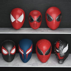 May include: A collection of seven superhero masks, primarily red, black, and blue. The masks feature various designs, including the iconic Spider-Man mask, and other character-inspired designs. The masks are displayed on shelves against a dark background.