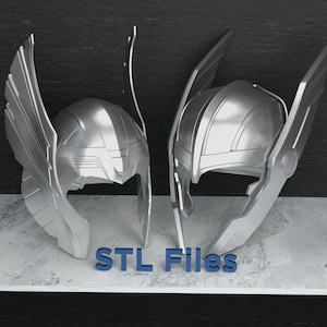 May include: Two silver-colored helmets with winged designs, reminiscent of a superhero's headgear. The helmets are displayed side-by-side, showcasing their intricate details. The text "STL Files" is visible in blue at the base.