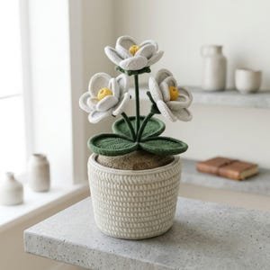 May include: A decorative potted plant with three white felt flowers and yellow centres. The flowers are on green stems and leaves, in a beige woven pot. The pot is on a grey surface.