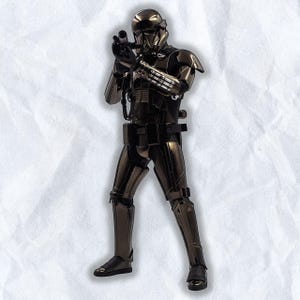 Puede incluir: Una figura de acción negra metálica de un soldado con un traje futurista, sosteniendo un rifle bláster. La figura tiene un casco, armadura y un cinturón de herramientas. La figura posa sobre un fondo blanco texturizado.