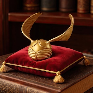 Peut inclure: Un vif-or doré aux ailes déployées est posé sur un coussin de velours rouge avec des bordures et des glands dorés. Le coussin est placé sur une pile de livres anciens, créant une ambiance vintage. Une bibliothèque en bois est en arrière-plan.