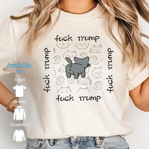 Op de afbeelding: Crèmekleurig T-shirt met de tekst "fuck trump" rond een grijze kattenillustratie. Het shirt is verkrijgbaar als t-shirt, hoodie en sweatshirt.