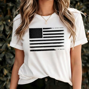 Può includere: T-shirt bianca con un design a bandiera americana nera invecchiata. La grafica della bandiera include il testo "the United States" e "of America". La maglietta ha maniche corte e girocollo.