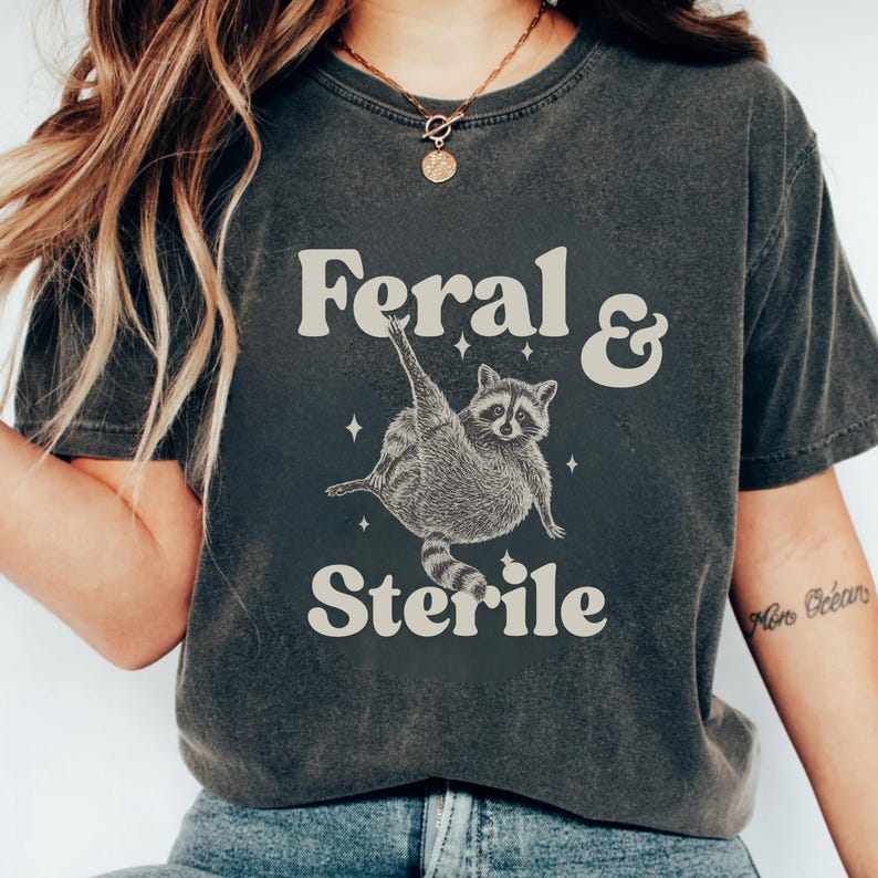 May include: Dark gray t-shirt with the words "Feral & Sterile" above and below a raccoon illustration. The raccoon is depicted in a playful pose. The shirt has a vintage, worn look.