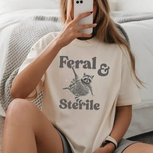 May include: Cream-colored t-shirt with the text "Feral & Sterile" in gray. A detailed illustration of a raccoon is featured between the words. The raccoon is depicted with its arms raised. The shirt is a casual, unisex style.