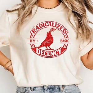 Op de afbeelding: Crèmekleurig T-shirt met een rode afbeelding. De afbeelding toont een duif met een takje in een cirkelvormig ontwerp met de tekst "RADICALIZED BY BASIC DECENCY".