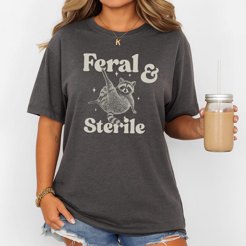 May include: Dark gray t-shirt with the text "Feral & Sterile" and a raccoon graphic. The raccoon is depicted in a playful pose. The shirt is a casual style, suitable for everyday wear.