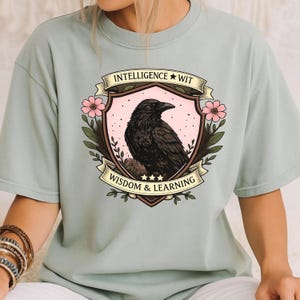 May include: A light green t-shirt featuring a crest with a black raven illustration. The crest is surrounded by pink flowers and green leaves, with the words "INTELLIGENCE WIT" and "WISDOM & LEARNING" on banners.