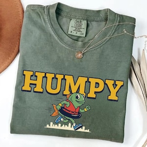 May include: Olive green t-shirt with the word "HUMPY" in yellow and navy blue block letters. A cartoon fish wearing a life preserver and running is printed below the word. A gold necklace is draped across the shirt.