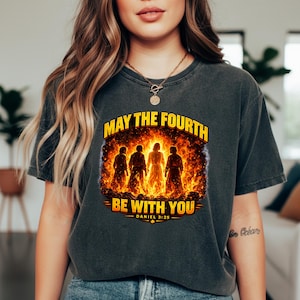May include: Dark grey t-shirt with the text "MAY THE FOURTH BE WITH YOU" in yellow. The graphic shows four silhouetted figures against a fiery backdrop. The text "DANIEL 3:25" is below the main text.