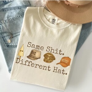 May include: Cream-colored t-shirt with the text "Same Shit. Different Hat." and illustrations of different hats. A tan hat and blue jeans are in the background.