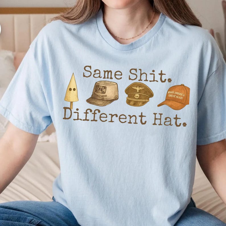 May include: Light blue t-shirt with the text "Same Shit. Different Hat." and illustrations of a pointed hat, a military cap, a military hat, and a "Make America Great Again" hat. The shirt is a crew neck.