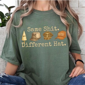 May include: Green t-shirt with the text "Same Shit. Different Hat." and illustrations of a KKK hood, a military cap, and a "Make America Great Again" hat. The shirt is a muted green color.