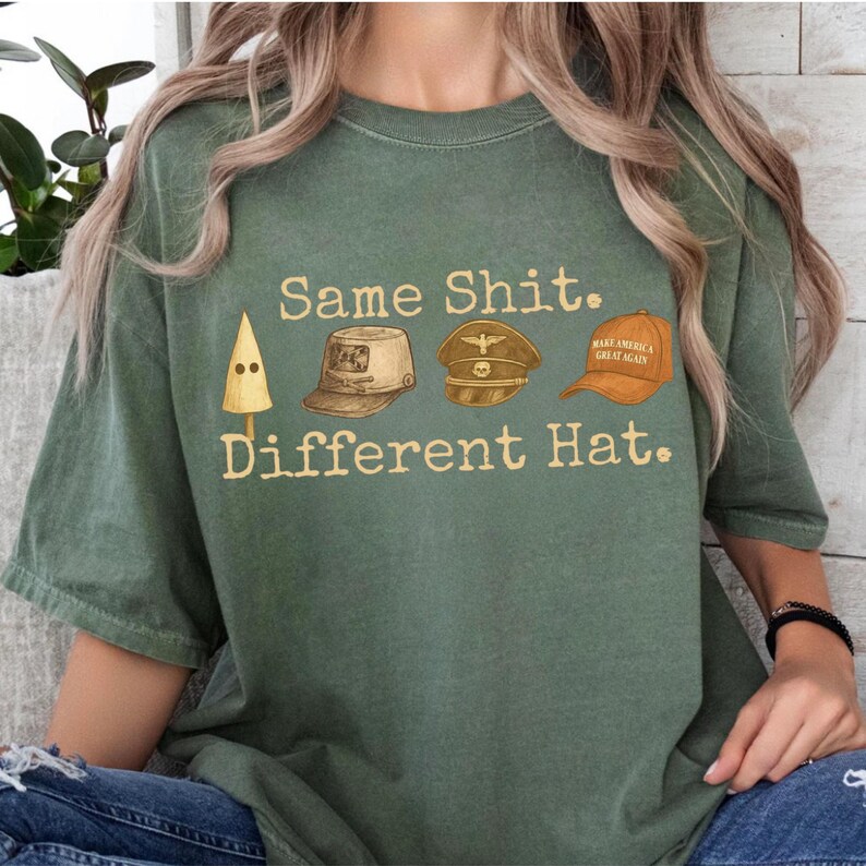 May include: Green t-shirt with the text "Same Shit. Different Hat." and illustrations of a KKK hood, a military cap, and a "Make America Great Again" hat. The shirt is a muted green color.