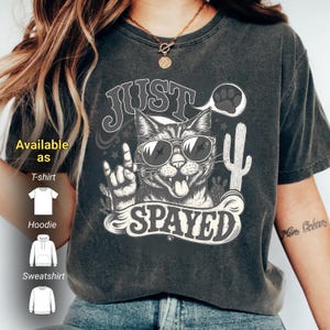 Puede incluir: Camiseta gris oscuro con un gráfico de un gato con gafas de sol haciendo un gesto de rock and roll. El texto dice "JUST SPAYED" con un bocadillo y una huella de pata. Disponible como camiseta, sudadera con capucha y sudadera.