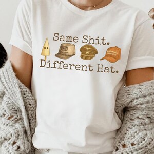 May include: White t-shirt with the text "Same Shit. Different Hat." and illustrations of different hats. The hats include a KKK hood, a military cap, and a "Make America Great Again" hat.