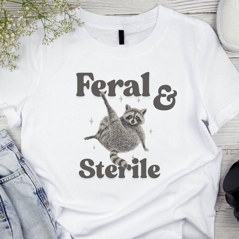 May include: White t-shirt with the words "Feral & Sterile" in gray lettering. A detailed illustration of a raccoon in a playful pose is centered below the text. The raccoon is gray and white with a striped tail.