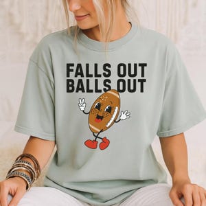 May include: A light green t-shirt with the text "FALLS OUT BALLS OUT" in black. The shirt features a cartoon football character with a smiling face, white gloves, and red shoes. The football is brown with white laces.
