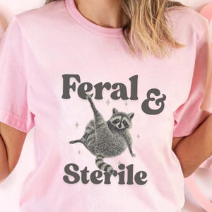 May include: A light pink t-shirt featuring the text "Feral & Sterile" in dark gray letters. A detailed illustration of a raccoon is centered below the text, with its arms raised. The shirt is a crew neck style.