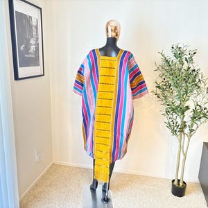 Pu&ograve; includere: Un abito tunica a righe colorate con scollo quadrato. L'abito presenta strisce verticali nei toni del rosa, blu, giallo e marrone. Un lungo pannello giallo con frange pende sul davanti. L'abito &egrave; esposto su un manichino.