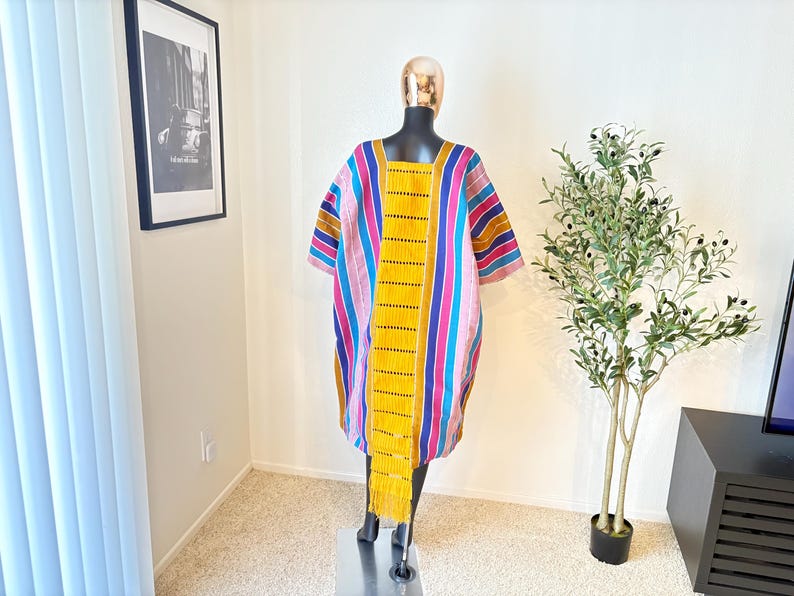 Pu&ograve; includere: Un abito tunica a righe colorate con scollo quadrato. L'abito presenta strisce verticali nei toni del rosa, blu, giallo e marrone. Un lungo pannello giallo con frange pende sul davanti. L'abito &egrave; esposto su un manichino.