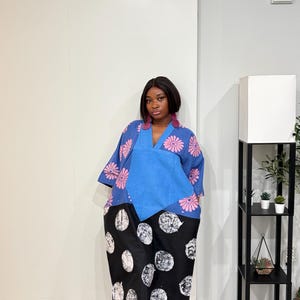 K&ouml;nnte beinhalten: Ein knielanges Kleid mit einem blauen V-Ausschnitt-Oberteil mit rosa Blumenmustern und einem einfarbigen blauen Mittelteil. Der untere Teil ist schwarz mit einem wei&szlig;en, kreisf&ouml;rmigen Punktemuster. Das Kleid wird mit wei&szlig;en Sandalen kombiniert.