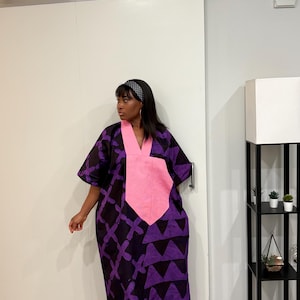 K&ouml;nnte beinhalten: Ein weit geschnittenes, bodenlanges Kaftankleid mit einem dunkelvioletten und schwarzen geometrischen Muster sowie einem markanten, einfarbigen rosa V-Ausschnitt-Einsatz. Das Kleid hat kurze, weite &Auml;rmel und eine lockere Passform, kombiniert mit wei&szlig;en Sandalen.